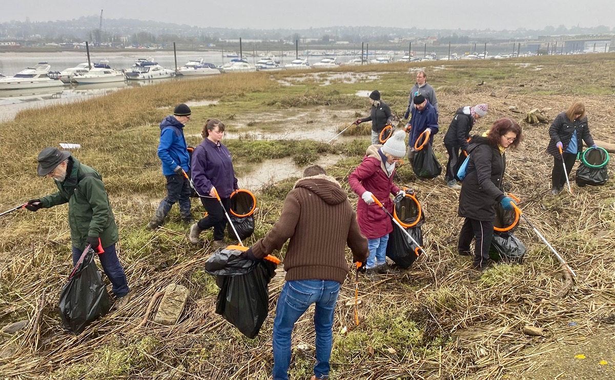 A group of about ten volunteers carrying bags and litter pickers remove flotsam and jetsam from the marshy foreshore of the River Medway